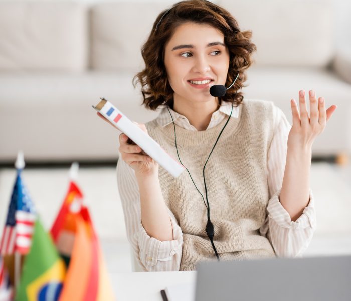 happy language teacher in headset holding French dictionary while talking during online lesson near international flags